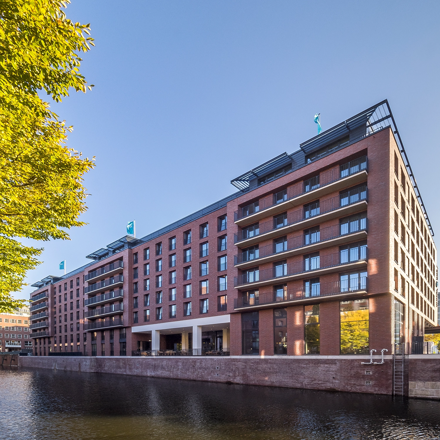 Das Bild zeigt das Motel One in der Admiralstraße in Hamburg, das direkt an einem kleinen Fluss liegt. Das Gebäude ist in rote Klinker gehüllt, was ihm eine markante, traditionelle Ausstrahlung verleiht. Die Sonne strahlt am blauen Himmel und wirft ihre warmen Strahlen auf das Motel. Besonders hervorzuheben sind die Absturzsicherungen des Geländersystems SIMPLUM von Abel Metallsysteme, die modern und funktional in das Gesamtbild integriert sind.