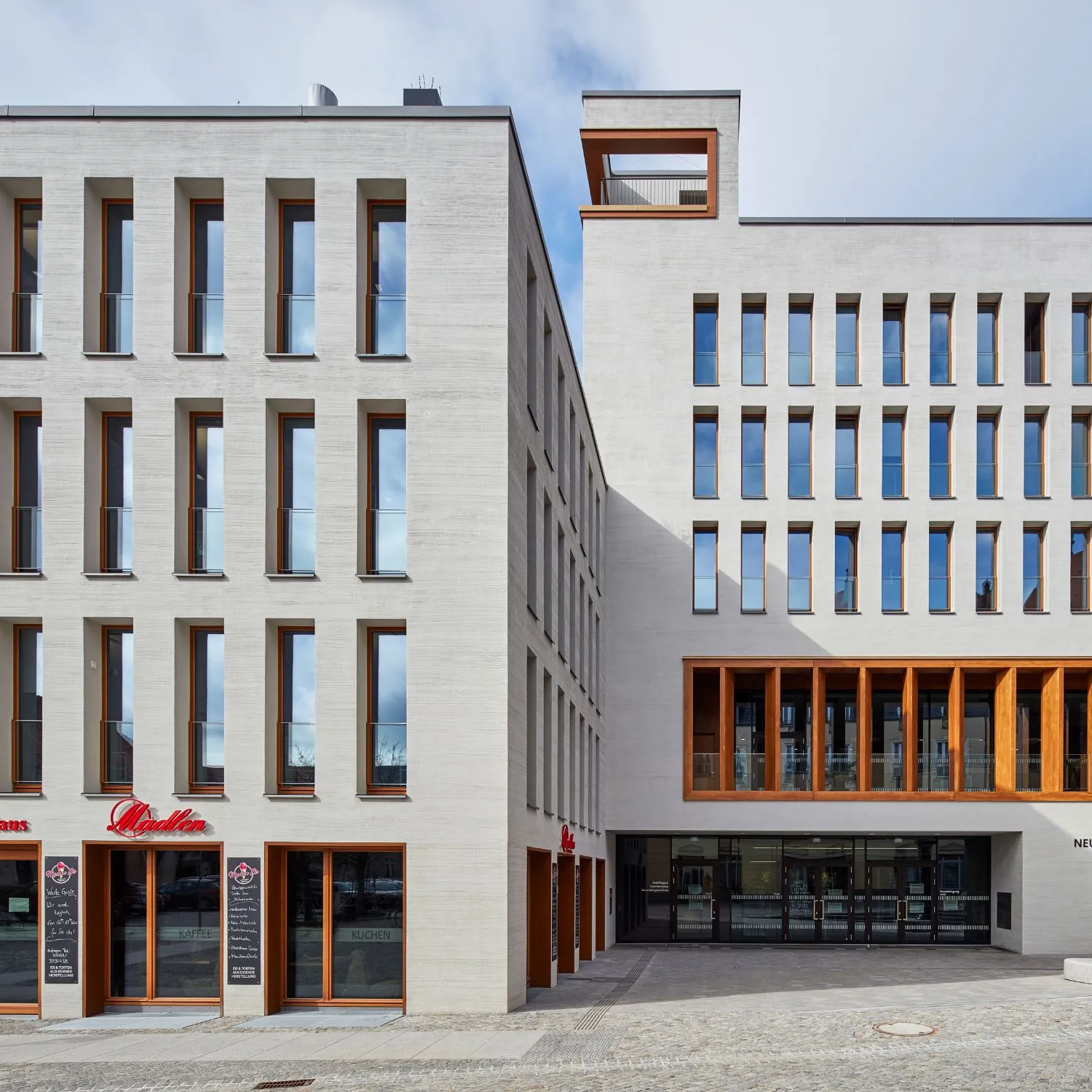 Das moderne Rathaus in Bernau zeichnet sich durch eine klare, strukturierte Fassade aus, die in hellem, sandfarbenem Stein gestaltet ist. Schmale, hohe Fenster gliedern die Fassade rhythmisch und werden durch die dezente Glasabsturzsicherung VITRUM von Abel ergänzt, die sich nahtlos in die Architektur einfügt. Die warmen Holzrahmen der Fenster setzen einen eleganten Kontrast zur Steinverkleidung und verleihen dem Gebäude eine moderne, einladende Note.